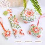 Cuatro piezas bordadas a mano siguiendo los patrones de punto de cruz de la colección Caléndula: un biscornú, un marcapáginas, una etiqueta y un colgante decorativo para tijeras, con un diseño floral vibrante en tonos naranjas y verdes, inspirado en las caléndulas. Texto indicando que es un producto digital con instrucciones disponibles en inglés, francés y español.