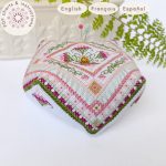Alfiletero realizado siguiendo el patron de punto de cruz para el Biscornú Tulipanes. La imagen muestra un pequeño cojín octogonal bordado con motivos geométricos y florales en varios tonos de rosa, burdeos y verde. Texto indicando que es un producto digital disponible en español, inglés y francés.