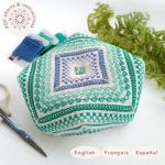 Patrón de punto de cruz para Mar Tropical. Alfiletero biscornu bordado a mano en tonos verdes, turquesa y azul, con diseño geométrico centrado en un cuadrado decorativo. Está rodeado de hilos, tijeras de bordado y una ramita de helecho. Texto indicando que es un producto digital disponible en español, inglés y francés