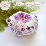 Alfiletero realizado siguiendo el patrón de punto de cruz Biscornú Violetas. Este pequeño cojín octogonal está bordado con motivos florales en varios tonos de violeta y verde con una gran violeta central rodeada de ramas con flores más pequeñas. Texto que indica que es un producto digital disponible en inglés, francés y español.