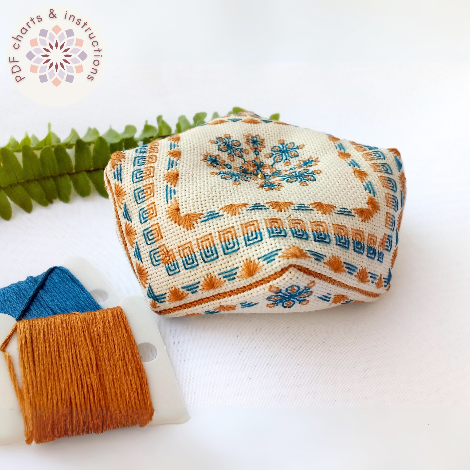 Copper-and-Teal-Biscornu-Back-stitch-Charts-and-Instructions Alfiletero realizado con el patrón de blackwork para el Biscornú Cobre y Azul. Pequeño cojín octogonal bordado en blackwork con motivos geométricos en tonos cobre y verde azulado. Bordado principalmente con puntadas lineales. El biscornú está presentado junto a cartones de hilos con tonos a juego y una hoja de helecho verde brillante. Texto indicando que se trata de un producto digital.