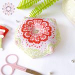 op view of the May Garden Biscornu cross stitch pattern, showcasing an octagonal pincushion richly embroidered with a large stylized floral motif in vibrant coral and pink tones. The design is framed by pale green geometric accents, stitched on white fabric. It is displayed on a white cloth, next to three thread bobbins in coordinating colors, a pair of pastel pink embroidery scissors, and a silver bead embellishment. Text indicating it is a digital product.