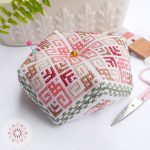 Hand-stitched cross stitch biscornu pincushion with a geometric rose-inspired pattern in soft pink, red, beige, and green tones, photographed with embroidery scissors and floss on a white background.