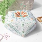 Dewdrop biscornu cross stitch pattern with an orange floral center, surrounded by soft green details, beads, and embroidery tools nearby.