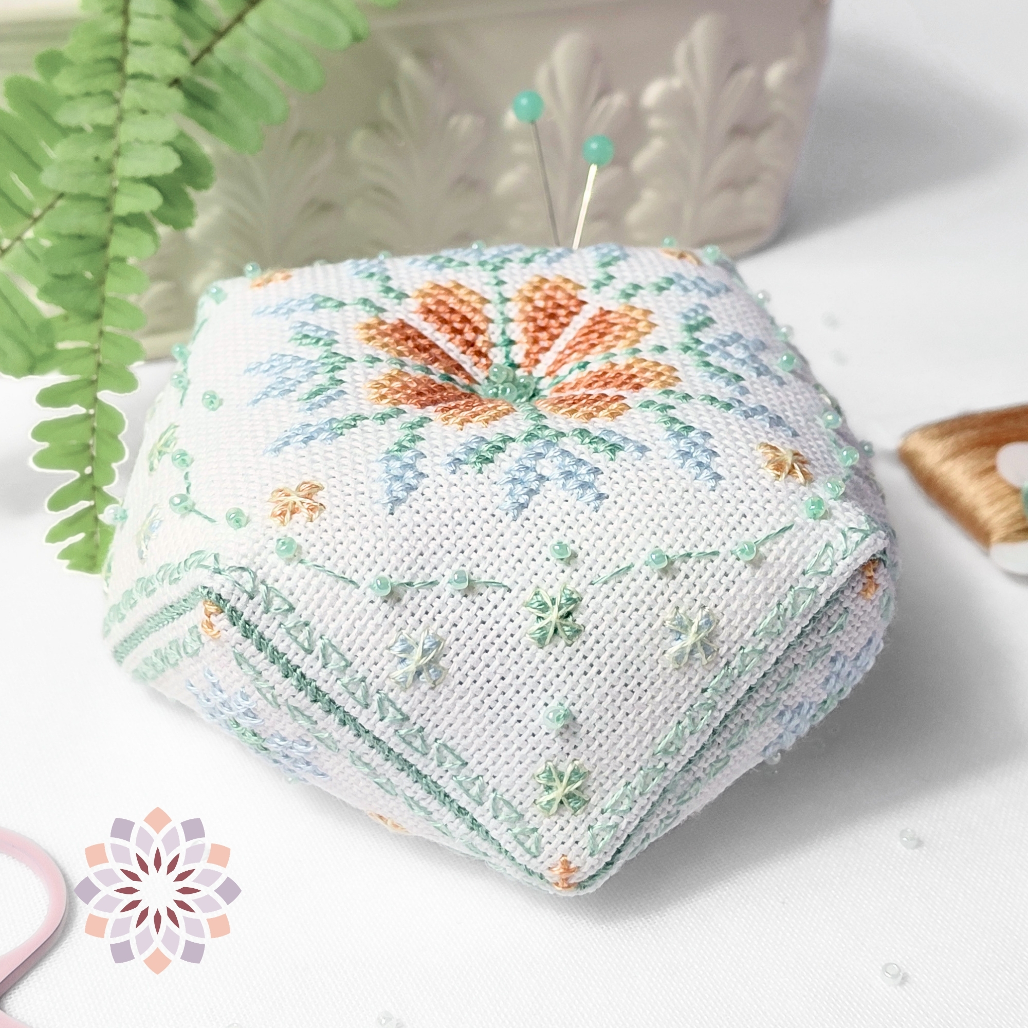Dewdrop-Biscornu-cross-stitch-pattern-No-badge Dewdrop biscornu cross stitch pattern with an orange floral center, surrounded by soft green details, beads, and embroidery tools nearby.