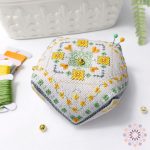 Golden Orchard floral biscornu cross stitch pattern finished as a decorative pincushion, embroidered with yellow flowers and green details on white fabric with a gold button.